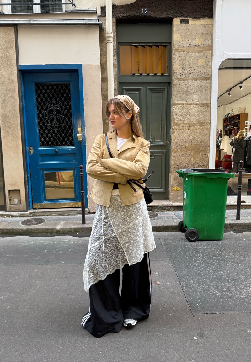 Jachetă din piele întoarsă bej peste o cămașă albă, combinată cu o fustă din dantelă. Pantaloni largi negri cu dungi albe și pantofi sport. În fundal se văd uși și un coș verde.