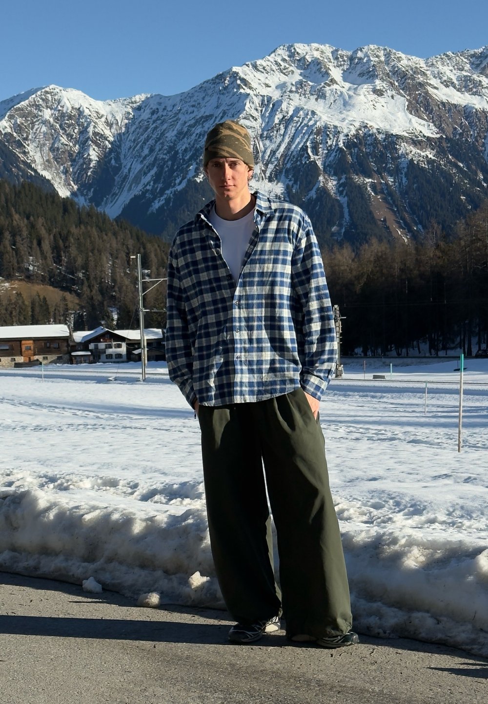Chemise en flanelle à carreaux bleus et blancs, portée sur un t-shirt blanc. Pantalon ample vert olive et bonnet camouflage. Arrière-plan de montagne enneigée.