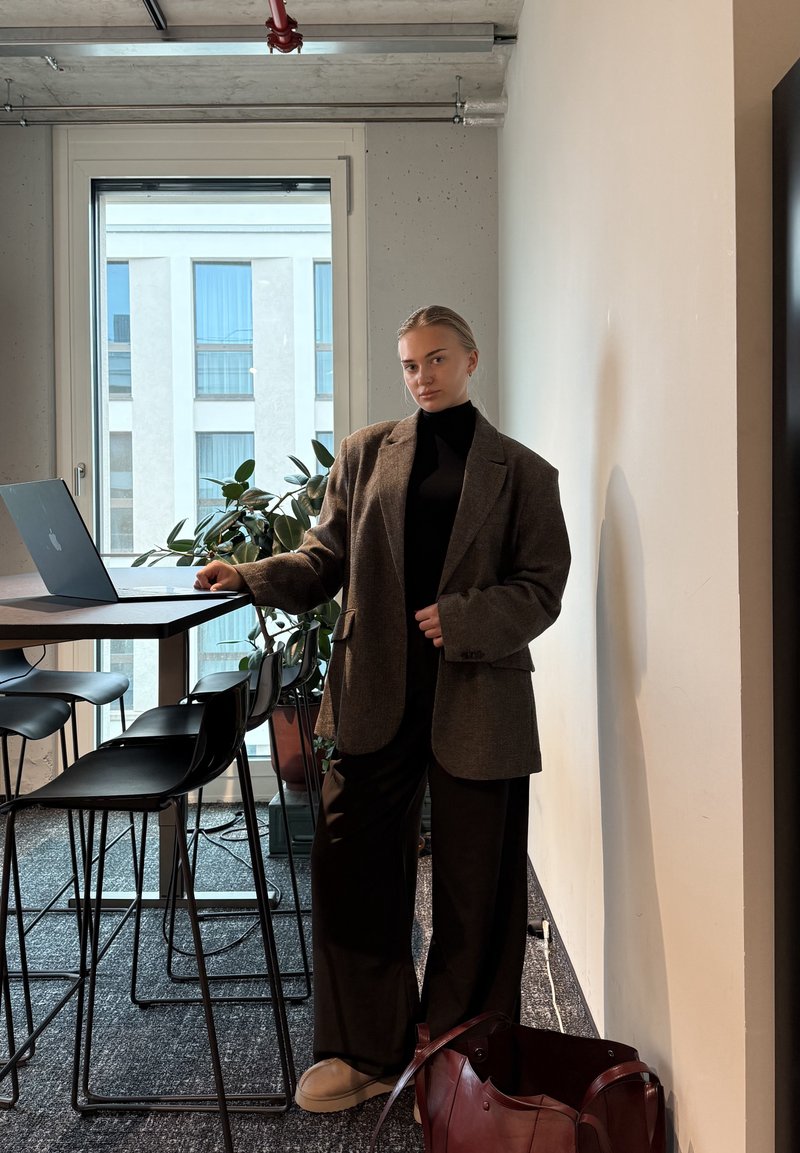 Brun oversized blazer over en sort turtleneck, parret med løse sorte bukser og beige sko. En rød taske ligger på gulvet ved siden af stolen.