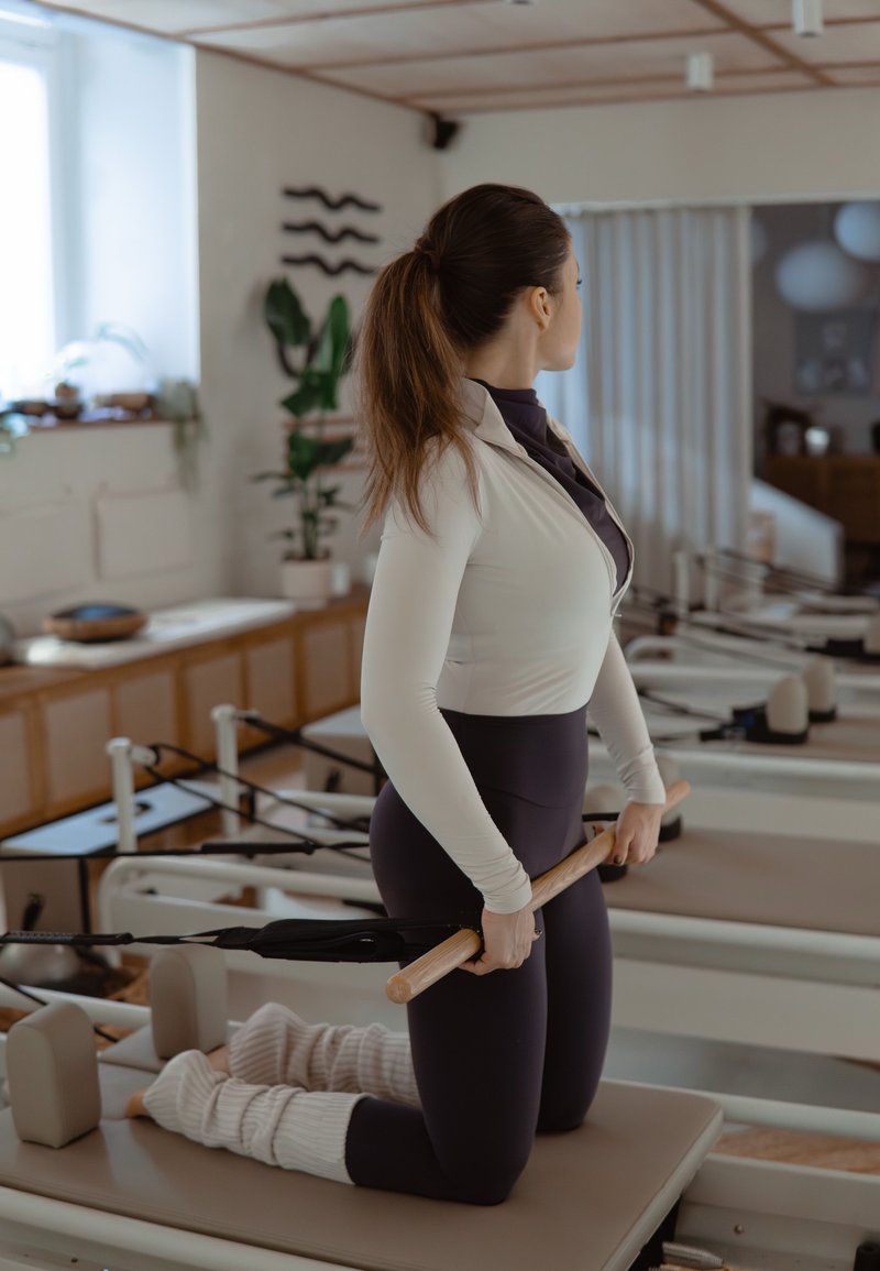 Kvinna i träningskläder som knäböjer på en Pilates-reformer och håller i en trästång med motståndsband i en ljus träningsstudio.
