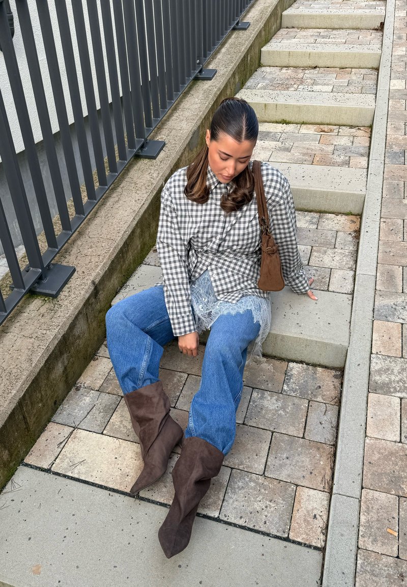 Jeune femme portant des bottes marron, un jean bleu, une chemise à carreaux et un sac à bandoulière marron, assise sur des marches en pierre extérieures à côté d'une rambarde en métal.