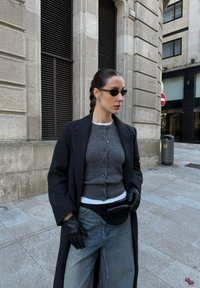 Femme portant des lunettes de soleil foncées, des gants noirs, un cardigan gris, un long manteau sombre et un jean, debout sur un trottoir urbain près d'un bâtiment en pierre.