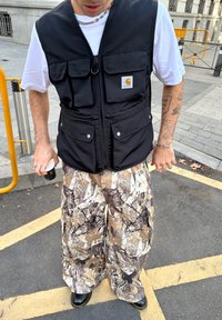 Osoba oblečená v čiernej vestě Carhartt, bielej tričku, maskárových cargo nohaviciach a čiernych čižmách, ktorá drží malý pohárik a stojí na ulici v meste.