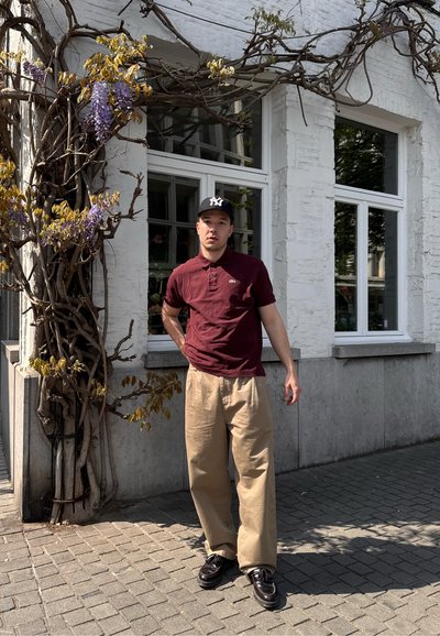  camiseta polo marrón, pantalones de pierna ancha color crema, gorra negra con logo blanco, zapatos negros, contra un fondo de pared blanca texturizada y vegetación.