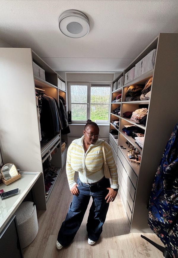 Žena so sklami a žltým pruhovaným sveterom pózuje v šatníkovej skrini s usporiadanými policami a zaveseným oblečením.