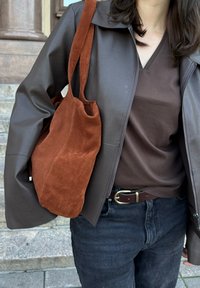 Καφέ δερμάτινο μπουφάν πάνω από καφέ μπλούζα V, συνδυασμένο με σκούρο τζιν και μια πορτοκαλί σουέτ τσάντα ώμου με απαλή υφή και μακριά λουριά.