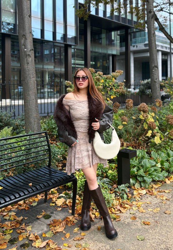 Vestido marrón claro sin hombros, chaqueta negra con acentos de piel, botas oscuras marrones hasta la rodilla y un bolso blanco curvado, rodeada de follaje otoñal.