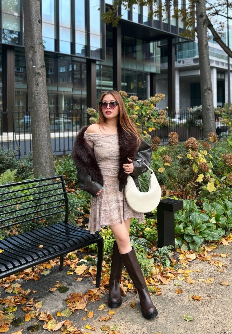 Heled pruun off-shoulder kleit, karvase äärega must jakk, põlveni ulatuvad tumesuunased saapad ja kumer valge käekott, ümbritsetud sügisel lehtedest.