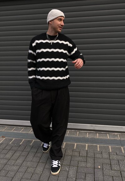 Suéter difuso en blanco y negro con un patrón ondulado, pantalones plisados negros y zapatillas negras con cordones blancos, todo sobre un fondo gris.