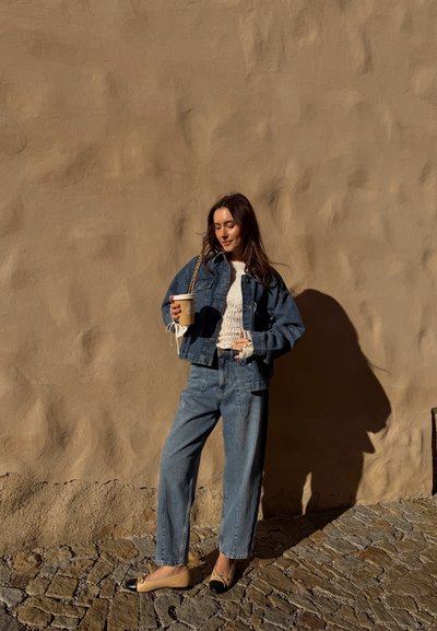 Mujer con chaqueta vaquera y vaqueros sosteniendo una taza de café, de pie frente a una pared beige texturizada con suelo de adoquines bajo la luz del sol.