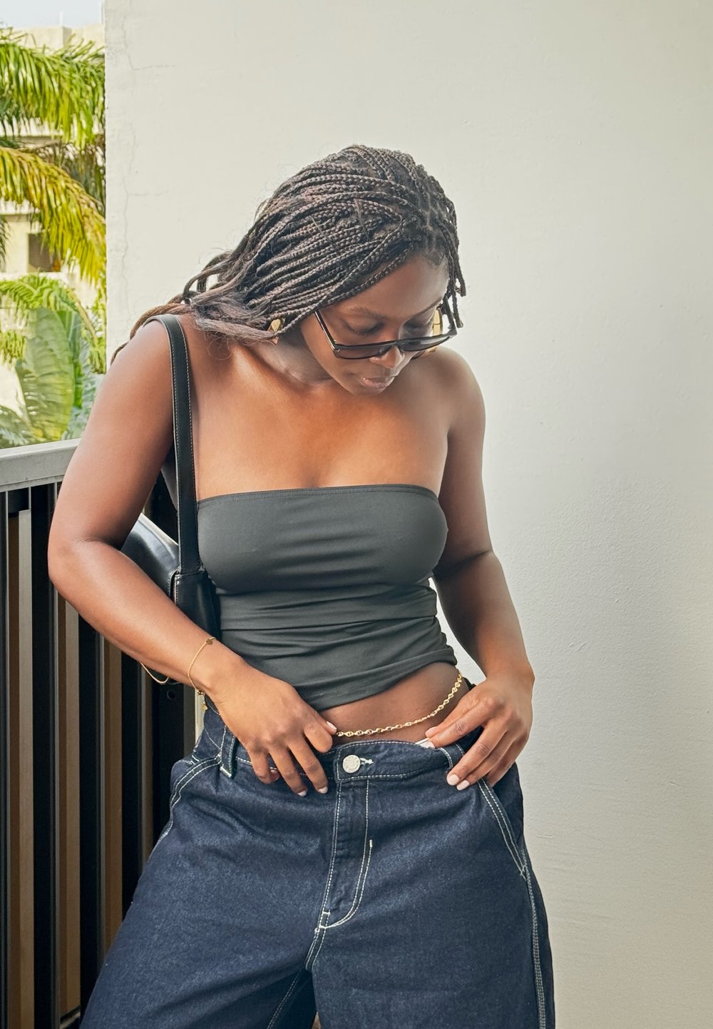 Top nero senza maniche, jeans in denim scuro a vita alta, catena dorata in evidenza in vita e borsa nera. Lo sfondo è caratterizzato da fogliame verde.