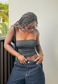 Top nero senza maniche, jeans in denim scuro a vita alta, catena dorata in evidenza in vita e borsa nera. Lo sfondo è caratterizzato da fogliame verde.