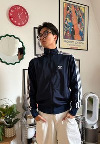 Jeune homme avec des lunettes portant une veste Adidas bleu marine et un pantalon blanc, debout dans une pièce avec des œuvres murales, une horloge rouge indiquant 12h55, et des plantes.