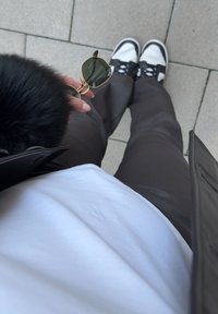 Czarne futrzane akcenty na ciemnej kurtce, biała koszula i brązowe spodnie. Okulary przeciwsłoneczne w złotej oprawie trzymane w ręku. Czarno-białe sneakersy na ziemi.
