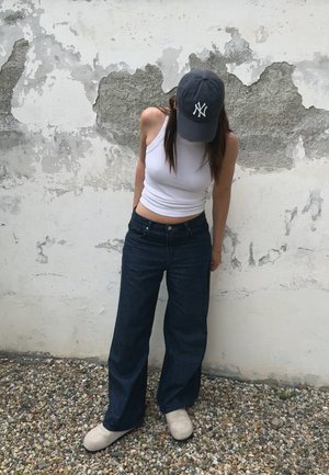 Jeans de mezclilla oscura de pierna ancha combinados con una camiseta sin mangas blanca ajustada y una gorra de béisbol gris. El look se completa con calzado de color crema.