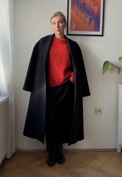 Persona joven que lleva un abrigo largo negro sobre un suéter rojo y una falda negra, de pie en el interior cerca de una planta y una obra de arte enmarcada en la pared.
