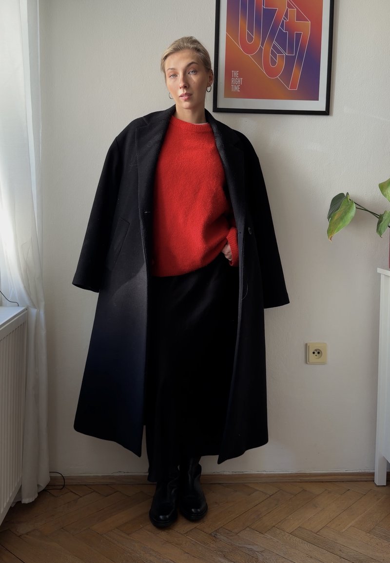 Jeune personne portant un long manteau noir par-dessus un pull rouge et une jupe noire, debout à l'intérieur près d'une plante et d'une œuvre d'art encadrée.