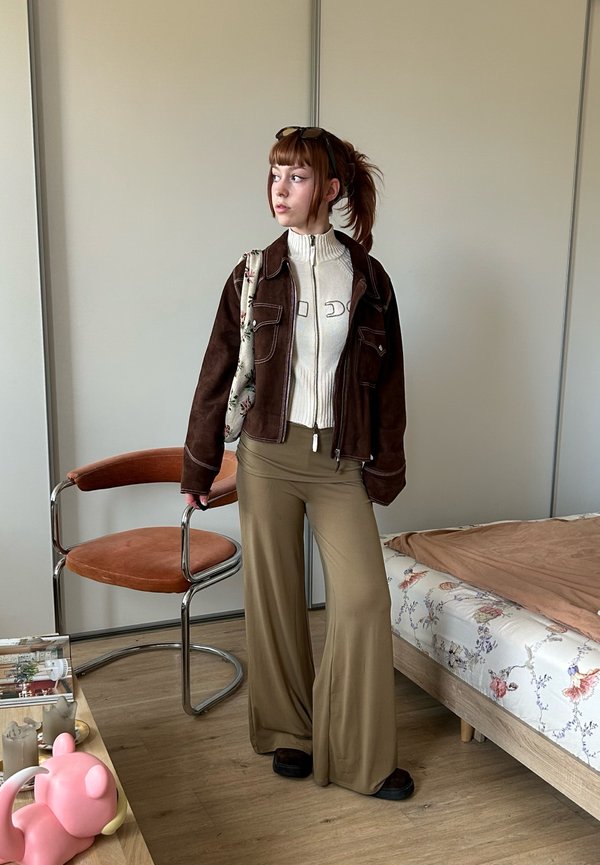 Joven mujer con cabello castaño en una coleta lleva chaqueta marrón, suéter blanco y pantalones anchos beige, de pie junto a una silla y una cama en una habitación.