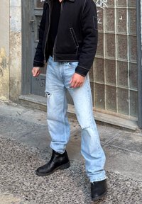 Uomo con giacca nera, jeans chiari strappati e stivali neri in piedi su un marciapiede testurizzato vicino a un muro in blocchi di vetro e una porta grigia.