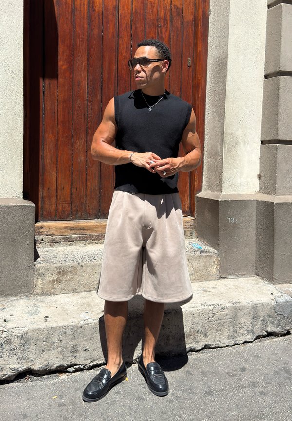 Man wearing black sleeveless top, beige wide shorts, black loafers, and sunglasses standing in front of a wooden door on a sunny day.