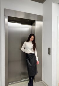 O tânără stă lângă un lift din oțel inoxidabil. Ea poart�ă un top de culoare deschisă, cu mâneci lungi și o fustă midi din piele neagră.
