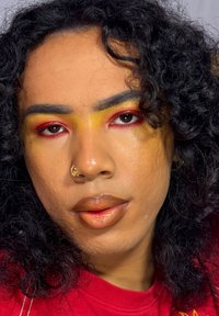 Gros plan sur un visage aux cheveux noirs bouclés, avec un fard à paupières orange-rouge, un dégradé jaune au-dessus des yeux, et des lèvres brunes brillantes. Anneau de nez visible.