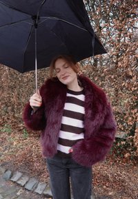 Jeune femme aux yeux fermés tenant un parapluie noir, portant un manteau en fausse fourrure bordeaux et un pull rayé, debout sur un sentier couvert de feuilles.