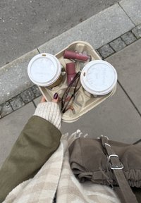 Kaksi kertakäyttökahvikuppia pahvitetarjottimessa, jossa on kaksi huulipunaa ja silmälasit, kädessä vihreää takkia ja vaaleaa neuletta käyttävä henkilö.