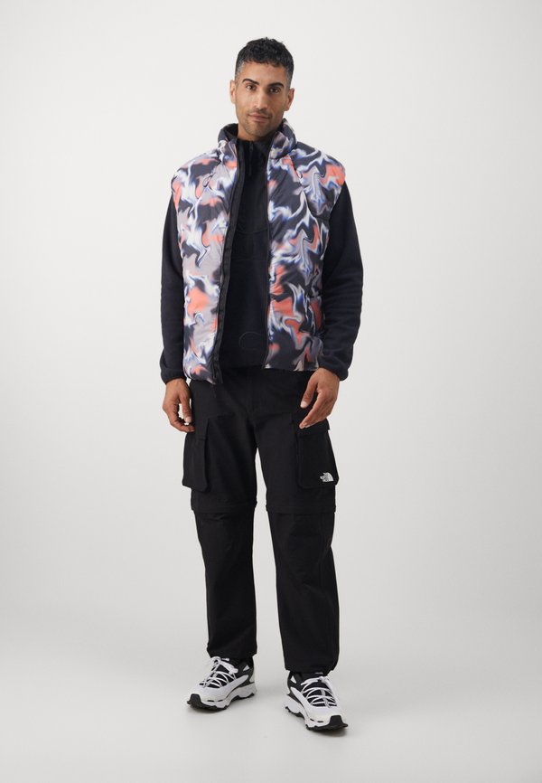 Farebný púderový vest s vírivým vzorom v modrej, oranžovej a bielej farbe; kombinovaný s čiernymi cargo nohavicami a bielymi teniskami.