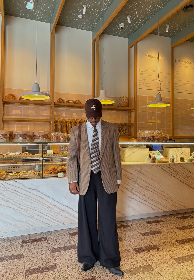 Homme avec une cravate à motifs, un blazer marron à carreaux, un pantalon noir large et une casquette se tient à l'intérieur d'une boulangerie avec du pain et des pâtisseries derrière un comptoir en verre.