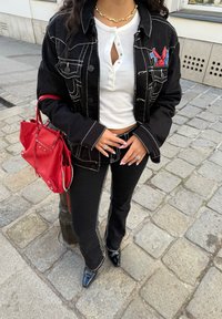 Jaqueta de ganga preta com costura branca, camiseta branca henley, calças de ganga pretas com riscas laterais, bolsa vermelha e sapatos pretos de bico.