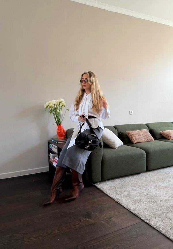 Femeie cu păr blond lung, purtând o bluză albă, fustă gri și cizme maro, stă pe o canapea verde ținând o poșetă neagră într-un living modern.