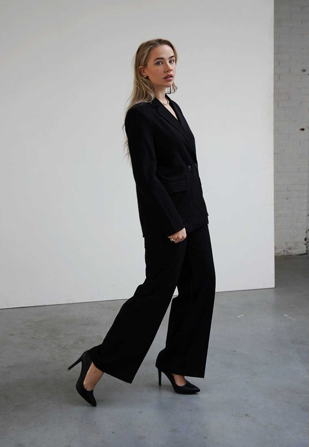 Fato preto sob medida com um blazer ajustado e calças de perna larga. Usado com sapatos de salto alto pretos. Tecido suave, detalhes mínimos e corte clássico.