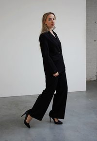 Fato preto sob medida com um blazer ajustado e calças de perna larga. Usado com sapatos de salto alto pretos. Tecido suave, detalhes mínimos e corte clássico.