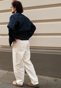 Giacca di jeans blu scuro, pantaloni bianchi larghi, scarpe marroni slip-on. Capelli ricci, in piedi di lato contro un muro grigio chiaro.