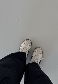 Valkoiset lenkkarit, joissa on kukkakoristeet ja mustat raidat, yhdistettyinä mustiin viivallisiin housuihin, harmaata lattiaa vasten.