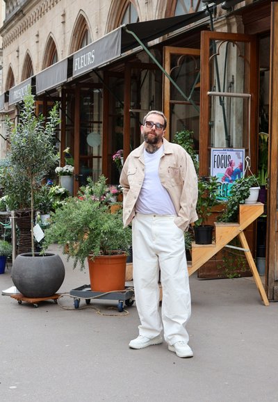 Chaqueta de pana beige sobre una camisa blanca, pantalones anchos blancos y zapatillas blancas. Rodeado de plantas en macetas y una vitrina de madera rústica.