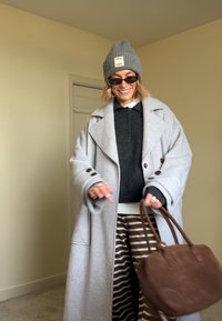 Grey wool coat with a double-breasted design, paired with a dark jumper and striped brown skirt. Holding a brown suede handbag.