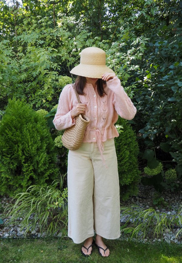 Vrouw die een roze trui, beige wijde broek, strohoed draagt en een gevlochten tas bij zich heeft in een groene tuinomgeving.