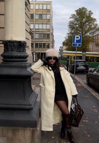 Cappotto bianco di lana con design doppiopetto, mini abito nero, stivali marroni sopra il ginocchio e un cappello leggero di pelliccia. Occhiali da sole neri e borsa inclusi.