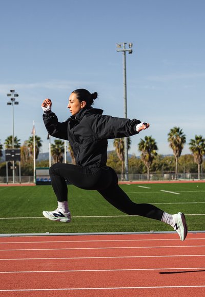 Cortavientos negro, mallas deportivas negras y zapatillas blancas con detalles. El sujeto está en pleno salto sobre una pista de atletismo con palmeras al fondo.