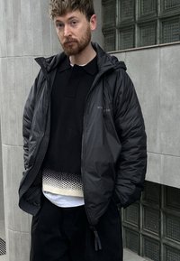 Svart dunjakke med hette, som har glidelåslukking og elastiske mansjetter, lagd over en svart polo og en mønstret strikket skjorte.