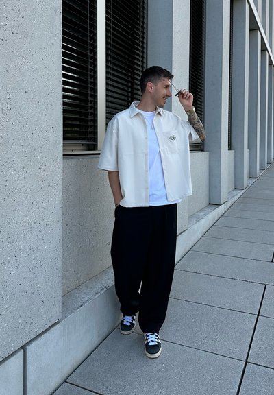 Camisa de manga corta color crema con un bolsillo en el pecho y cierre de botones, combinada con pantalones negros holgados y zapatillas oscuras con acentos blancos.