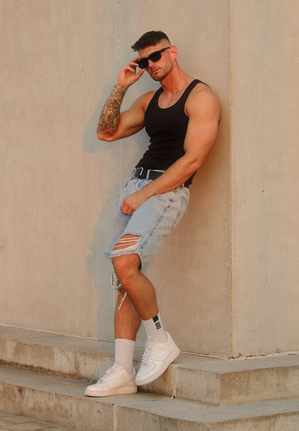 Črn top z naramnicami, uničene svetlo modre denim kratke hlače, bele superge in črna sončna očala. Mišičast telesni postavo, tetovaže na desni roki.