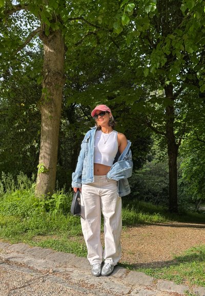 Chaqueta de denim, top corto blanco y pantalones holgados blancos; gorra rosa, zapatillas plateadas. Rodeada de vegetación, de pie en un camino de gravilla.