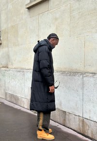 Črni puhasti plašč s kapuco in dolžino do tal. Oblečen z olivno zelenimi hlačami in rumenimi delovnimi škornji, stoji ob kamnitem zidu.