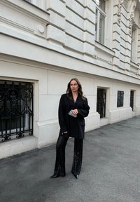 Fekete, oversize blézer, flitteres fekete harangnadrág, hegyes orrú fekete cipők, és egy csillogó ezüst kistáska egy világos színű épület háttérrel.