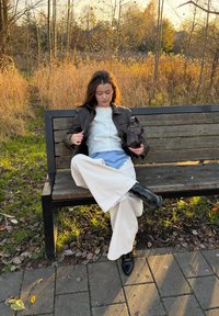 Una persona che indossa una giacca di pelle scura, pantaloni crema a gamba larga e stivaletti neri, seduta su una panchina di legno in un ambiente naturale.