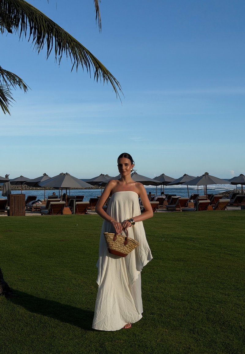 Mujer con vestido blanco sin tirantes sosteniendo un bolso tejido, de pie sobre la hierba cerca de sillas de playa y sombrillas con el océano al fondo.