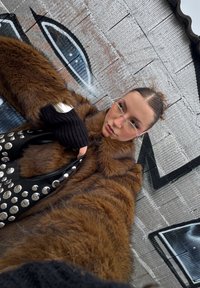 Καφέ ψεύτικο παλτό με φαρδιά μανίκια, μαύρη δερμάτινη τσάντα με ασημένια καρφιά και ριγέ μαύρα γάντια μπροστά σε υφή φόντου.